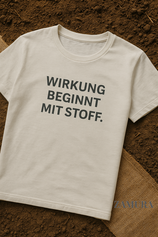 T-Shirt mit der Aufschrift „Wirkung beginnt mit Stoff“ liegt auf brauner Erde. Unten rechts ist das Logo der Marke ZAMURA gut sichtbar. Die Szene vermittelt Naturverbundenheit, Einfachheit und soziale Wirkung.